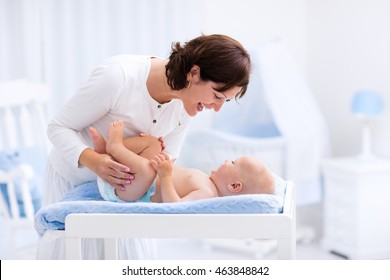 Young mother taking care of little boy in white sunny nursery with changing table, baby crib and rocking chair. Diaper change and clothing. Mom and son in beautiful bedroom at home.