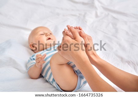 Young mother taking care of little baby son wearing bodysuit lying on bed at home looking joyful at mom doing pediatric massage holding his small feets