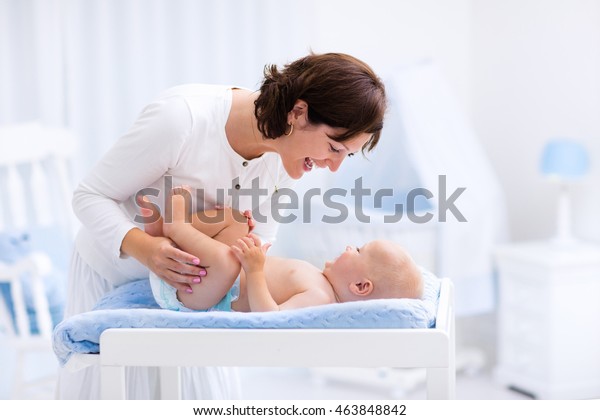 Young\
mother taking care of little boy in white sunny nursery with\
changing table, baby crib and rocking chair. Diaper change and\
clothing. Mom and son in beautiful bedroom at\
home.