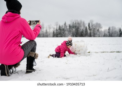 Young Mother Taking Photo With  Mobile Phone Of Her Toddler Child Playing In Snow Outside In Beautiful Winter Nature.