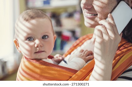 Young Mother Talking On A Phone Having Her Baby In A Carrier