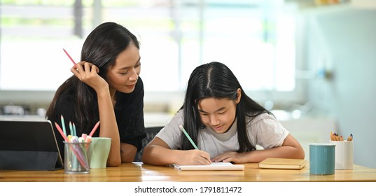 A young mother is teaching a daughter to do homework at the wooden table.