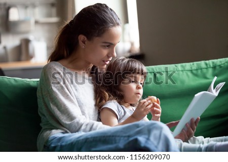 Young mother teaching daughter holding book sitting on sofa, mom or babysitter embracing little girl reading fairy tale to kid, nanny with child having fun together at home, family leisure activities