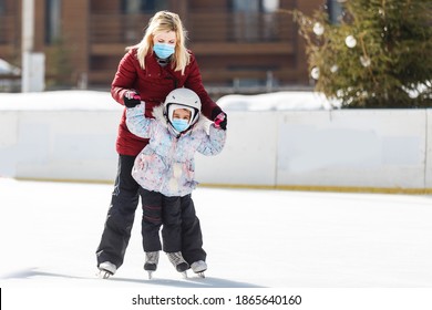 Young Mother Teaching Her Little Daughter Ice Skating At Outdoor Skating Rink. Family Wearing A Medical Mask During COVID-19 Coronavirus Enjoy Winter On Ice-rink Outdoors