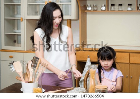Young mother is teaching how to cooking for her daughter in the kitchen. Cute woman and child holding pasta (spaghetti) teasing each and having fun.