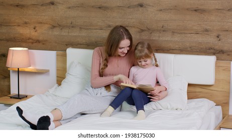 Young Mother And Teen Child Daughter Lying In Bed At Home Bedroom On Duvet Blanket And Reading Book Bedtime Stories And Fairytales Together With Warm Lamp At Night. Family Bonding, Parenthood Concept