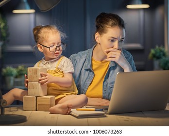 Young Mother With Toddler Child Working On The Computer From Home At Night. 