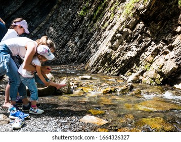 Young mother tries to help her daughter take off her shoes to wet her feet in the mountain stream during trekking on a sunny warm summer day. Concept of tourism and travel with children - Shutterstock ID 1664326327