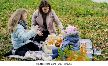 Young Mother And Two Little Daughters Are Drinking Tea At A Picnic In Nature.