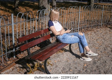 A Young Mother Is Waiting For Her Child On The Playground. Looks At Smartphone, Bored