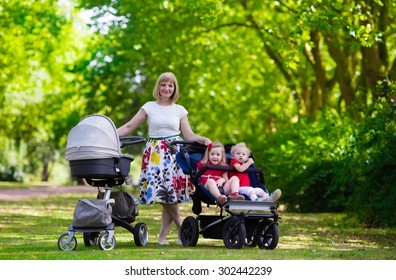 Young Mother Walking In A Park With Children In Pushchair. Mom And Kids In A Buggy Walk In Forest. Woman Pushing A Double Stroller For Twin Boy And Girl And Newborn Baby. Active Family Outdoors.