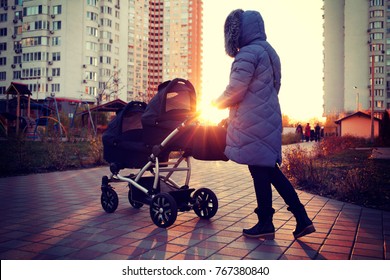 Young Mother Walking In A Park With Children In Pushchair. Mom And Kids In A Buggy Walk In Forest. Woman Pushing A Double Stroller For Twin Boy Newborn Baby. Active Family Outdoors.