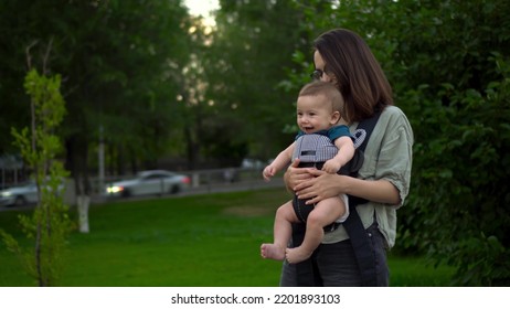 A young mother walks with a child sitting in a kangaroo backpack. Woman with glasses with a child in the park. The newborn baby laughs. Closeup. - Shutterstock ID 2201893103