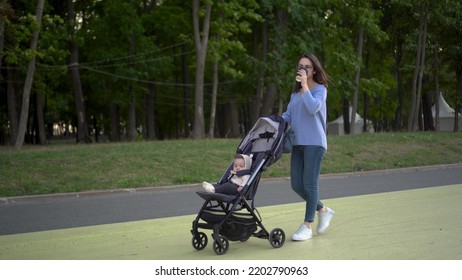 A young mother walks with a stroller in the park with glasses of coffee in her hand. A woman drinks coffee and carries her son in a stroller. - Shutterstock ID 2202790963