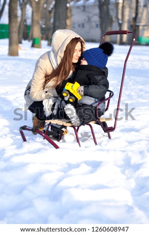 A young mother wants to kiss her little toddler son for a walk in a winter park on a sled.