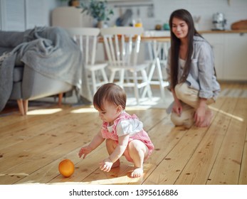 Young Mother Watching Her Cute Baby Daughter Playing Orange Fruit On Hardwood Floor At Home 