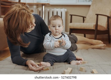 Young Mother Watching Over Her Cute Little Baby Boy As He Plays On The Carpet In The Living Room With Wooden Blocks In A Close Up Low Angle Portrait