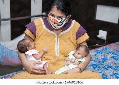 Young Mother Wearing Mask Sitting On Bed With Her Two Newborn Twin Babies, Stay Home Taking Extra Care Due To Covid-19 Pandemic. Indian Mother With Her Babies.