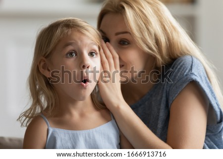 Young mother whisper in surprised cute little daughter ear tell secret, millennial mom or nanny play with small preschooler girl child, share close intimate moment at home, gossip or chat together