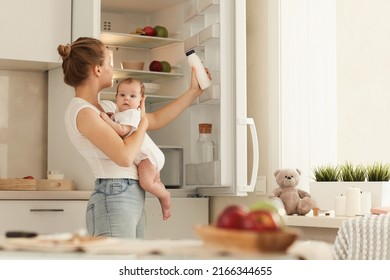 Young mother in white T-shirt and blue jeans with baby in her arms looking for bottle with baby formula in fridge