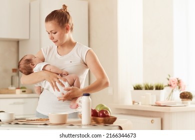 Young mother in white T-shirt rocking her baby in kitchen standing next to table covered with food - Powered by Shutterstock