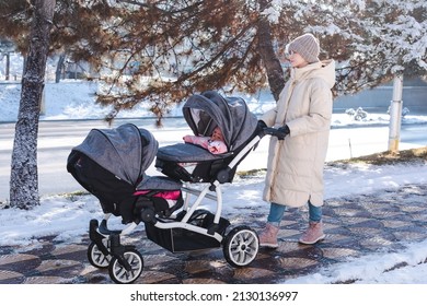 Young mother in winter clothes with double baby stroller for twins outside in the cold winter weather