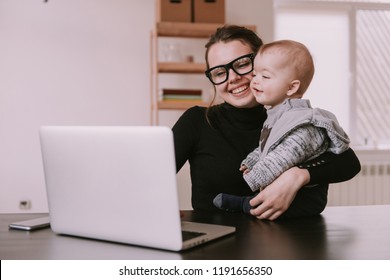 Young Mother Working At Home With Laptop Holding Her Little Son. Family, Mother Working With Child. Mom Is Using A Computer And Spending Time With Her Cute Baby Boy At Home. Son And Mummy