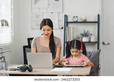 Young mother working on laptop beside her daughter playing on tablet at home office. - Powered by Shutterstock