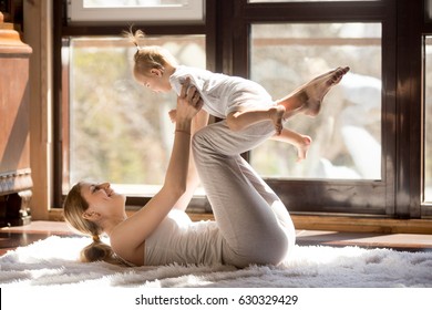 Young mother working out wearing white sportswear,  exercising at home with baby daughter, leg lifting with kid as a weigh, exercising and bonding with child, enjoyment. Healthy lifestyle concept 