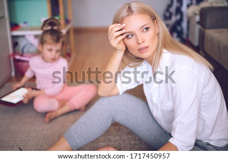 Young mother is worried and sad at home on the floor, the child is watching cartoons on the tablet and playing, a naughty child, family concept.