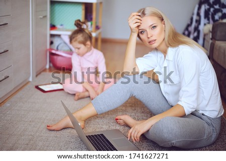 Young mother is worried and sad at home on the floor, the child is watching cartoons on the tablet and playing, a naughty child, family concept.