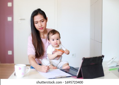 Young mother writing on paper work from home with her baby during coronavirus,Covid-19 pandemic quarantine. Social distancing lockdown isolation. Asian mommy working with tablet online meeting and kid