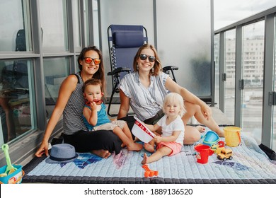 Young Mothers Spending Time Together With Children Babies On Balcony At Home. LGBTQ Female Parents Playing With Kids. Staycation During Coronavirus Covid-19 Pandemic In The World.