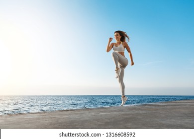 Young motivated happy sporty female runner fitness instructor, jumping, excercise near sea enjoying fresh morning air training, smiling facing sunlight, workout wearing activewear, running in quay - Powered by Shutterstock