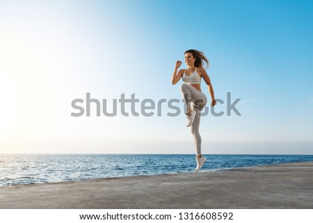 Young motivated happy sporty female runner fitness instructor, jumping, excercise near sea enjoying fresh morning air training, smiling facing sunlight, workout wearing activewear, running in quay