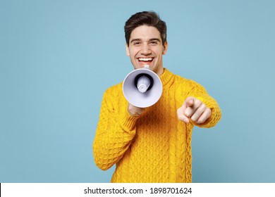 Young Motivating Leader Student Man 20s In Casual Knitted Yellow Fashionable Sweater Scream Aside Shout In Megaphone Point Index Finger Camera On You Isolated On Blue Color Background Studio Portrait