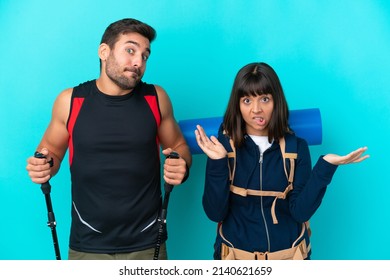 Young mountaineer couple with a big backpack isolated on blue background making unimportant gesture while lifting the shoulders