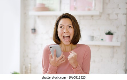 Young multi ethnic asian woman feel excited celebrating bid win success victory looking at smartphone, excited girl screaming yes winning online game app on cell phone sit on sofa at home