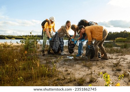 Young multi ethnic group of volunteers cleaning up the shore of a forest lake together, they are collecting trash and holding garbage bags. Concept of clean nature, ecology and sustainability. High