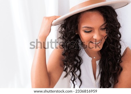 Young multi ethnic woman with long curly dark hair sitting on floor in casual neutral colors with straw hat