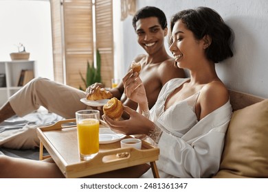 A young multicultural couple enjoys a cozy breakfast in bed, sharing laughter and a delicious meal. - Powered by Shutterstock