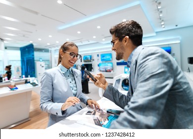 Young Multicultural Couple Looking For New Smart Phone To Buy. Woman Looking At Catalog While Man Holding Smart Phone.