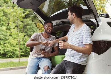 Young Multicultural Male Friends Sitting In Open Car Back, Drinking Coffee With Brofist Gesture