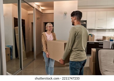 Young Multiethnic Couple Carrying Cardboard Box In New Apartment. Smiling Caucasian Girl And Middle Eastern Man Looking At Each Other. Concept Of Home Relocation. Interior Of Modern Spacious Flat