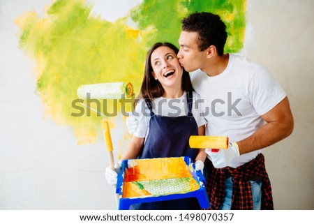 Young multiethnic couple decorate their new home. Husband and wife are painting the wall with rollers that are dipped in yellow and green paint. They are happy. Man gives girl kiss. Renovations home