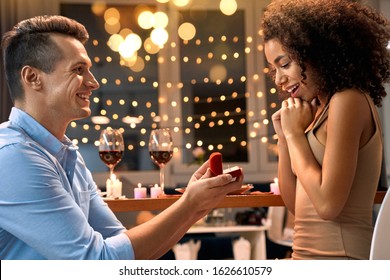 Young multiethnic couple having dinner romantic date at kitchen at home man standing on one knee holding box with proposal ring proposing to woman smiling surprised
