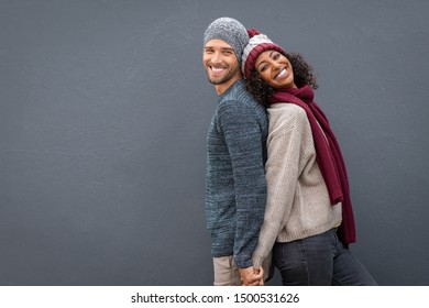 Young Multiethnic Couple Holding Hands While Standing And Leaning On Each Other Back. Happy Man And Black Woman Standing Back To Back And Smiling Against Grey Wall With Copy Space.