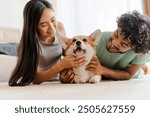 Young multiethnic couple is lying on a carpet and petting their corgi dog. They are both smiling and looking at their pet with love