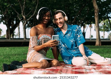 A young multiethnic couple with a transgender woman are spending time together outdoors, sitting on a blanket in a park while sharing headphones and looking at a smartphone - Powered by Shutterstock