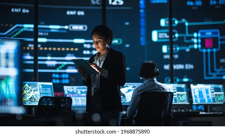 Young Multiethnic Female Government Employee Uses Tablet Computer In System Control Monitoring Center. In The Background Her Coworkers At Their Workspaces With Many Displays Showing Technical Data.